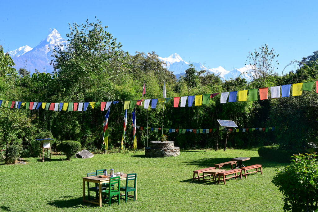 Sunny garden with colorful prayer flags at Kalsee Eco Lodge Nepalstrung across, green tables and benches, and snow-capped mountains in the distance.