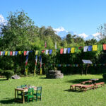 Sunny garden with colorful prayer flags at Kalsee Eco Lodge Nepalstrung across, green tables and benches, and snow-capped mountains in the distance.